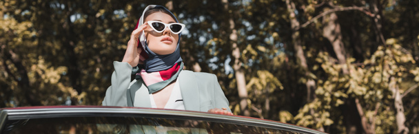 Elegante donna che tocca gli occhiali da sole vicino all'auto all'aperto, banner - Foto, immagini