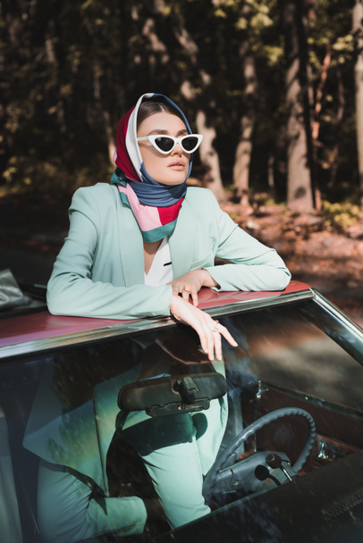 Stylish woman in sunglasses and headscarf looking away from vintage roofless car  - Foto, Imagem