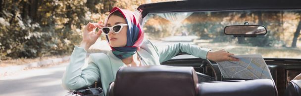 Mujer con mapa tocando gafas de sol en coche sin techo en primer plano borroso, bandera - Foto, Imagen