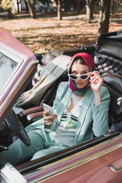 Woman in sunglasses holding smartphone and looking at camera in retro car on blurred foreground - 写真・画像