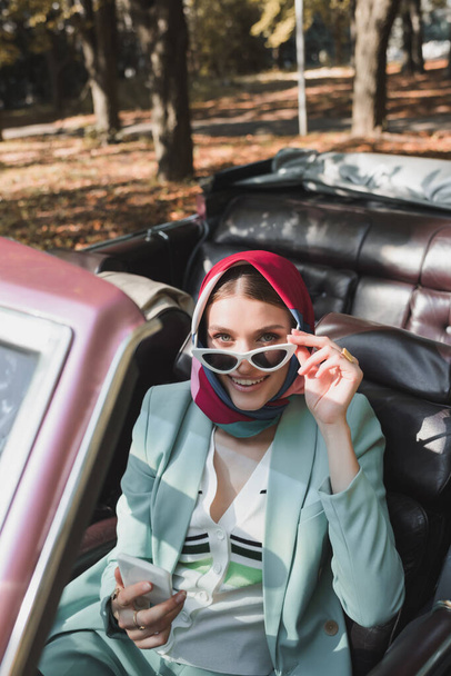 Woman holding sunglasses and smartphone in roofless car on blurred foreground outdoors  - Photo, Image