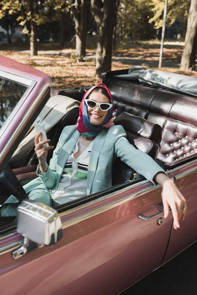 Positive stylish woman holding smartphone on driver seat of retro car on blurred foreground - 写真・画像