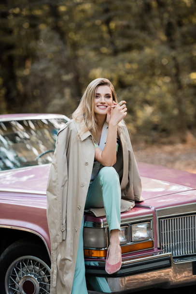 happy stylish woman looking at camera while sitting on hood of vintage cabriolet - Photo, Image