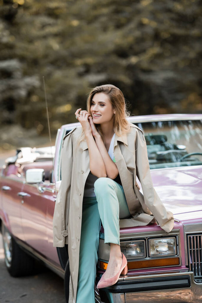 happy young woman in cape looking away while sitting on cabriolet hood on blurred background - Fotoğraf, Görsel