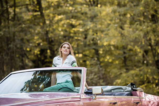 young stylish woman looking at camera while posing in vintage convertible car - 写真・画像