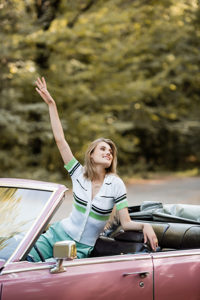 smiling woman looking away while sitting in convertible car with hand in air - Photo, Image