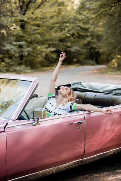 joyful woman relaxing in cabriolet while sitting with raised head, closed eyes and hand in air - 写真・画像