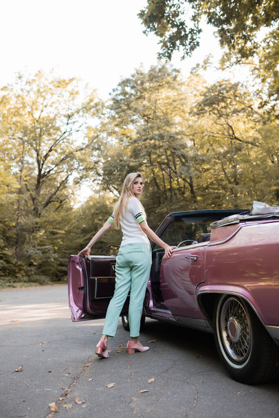 jonge stijlvolle vrouw kijken weg terwijl het openen van de deur van vintage cabriolet - Foto, afbeelding