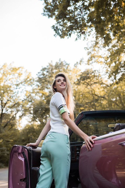 vrolijke jonge vrouw kijken weg tijdens het openen van de deur van vintage cabriolet - Foto, afbeelding