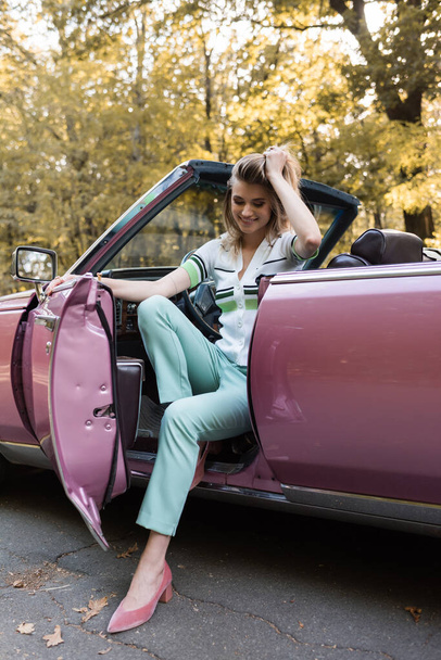 happy stylish woman touching hair while stepping out cabriolet - Photo, Image