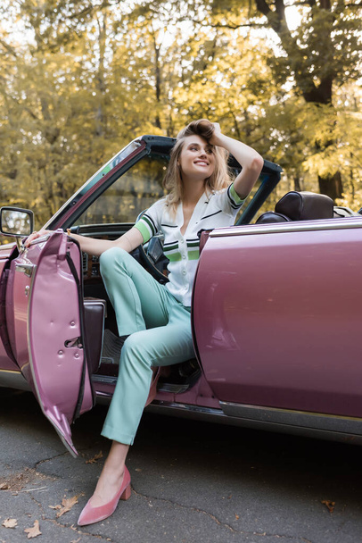 cheerful woman looking away and touching hair while stepping out cabriolet - Foto, Imagen