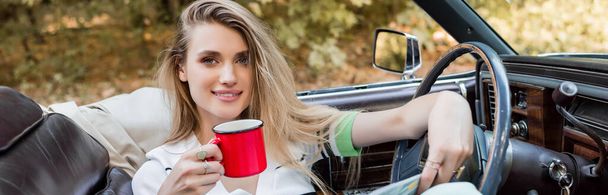 szczęśliwa młoda kobieta uśmiechnięta do kamery trzymając filiżankę kawy w samochodzie z kabrioletem, baner - Zdjęcie, obraz