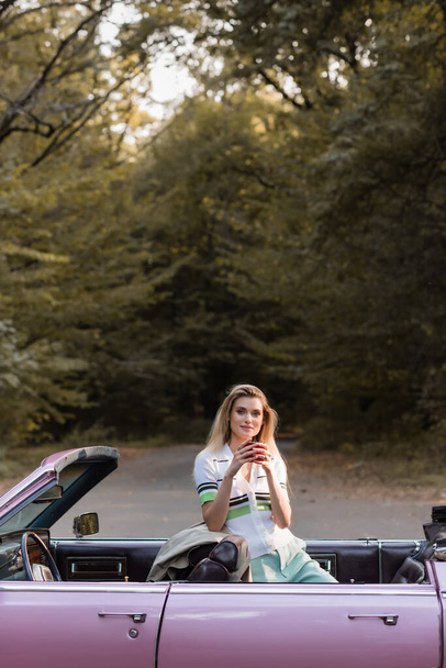 νεαρή γυναίκα κοιτάζοντας κάμερα κρατώντας ένα φλιτζάνι καφέ σε cabriolet στο δρόμο κοντά στο δάσος - Φωτογραφία, εικόνα