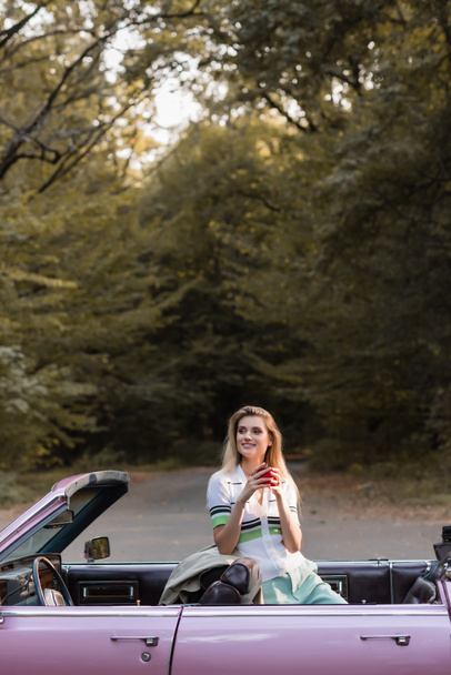 sonriente mujer sosteniendo taza de café y mirando lejos de cabriolet vintage cerca del bosque - Foto, imagen