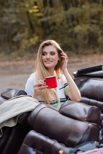 happy woman looking away and touching hair while holding cup of coffee in cabriolet on blurred foreground - Fotó, kép