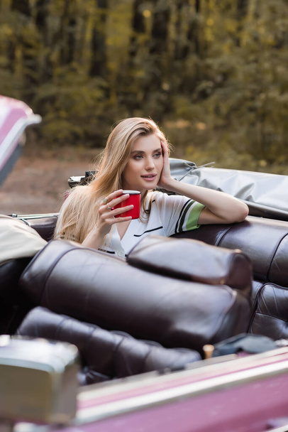 smiling woman holding hand near face and looking away while sitting in cabriolet with cup of coffee on blurred foreground - Foto, Bild