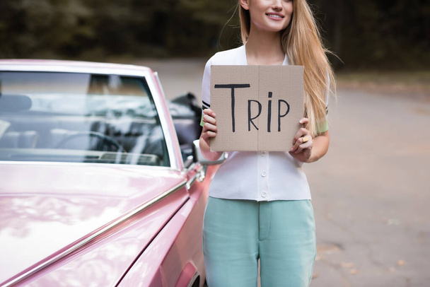 partial view of young woman holding placard with trip lettering near vintage cabriolet on blurred background - Фото, изображение