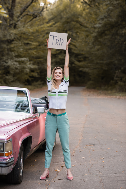 joven mujer sonriente mirando a la cámara mientras sostiene pancarta con letras de viaje cerca de cabrio vintage  - Foto, Imagen