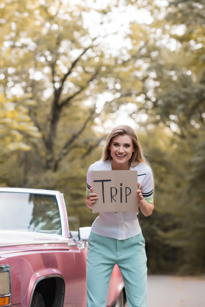 excited young woman holding placard with trip lettering near retro convertible car - Fotoğraf, Görsel