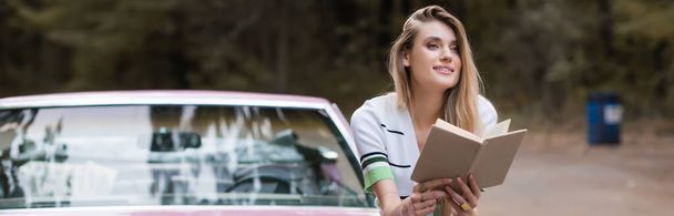 femme souriante assise sur la voiture avec le livre et regardant loin sur fond flou, bannière - Photo, image