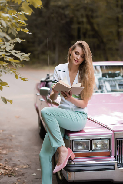 stylish woman reading book while sitting on hood of vintage cabriolet - Foto, Imagem