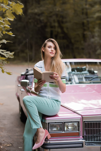 young woman looking away while sitting on hood of retro cabriolet and holding book on blurred background - Foto, Imagem