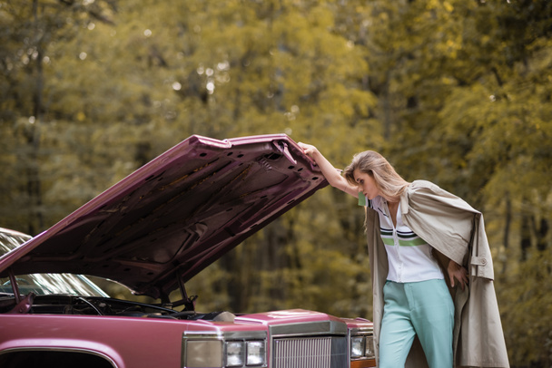 mladá žena v plášti při pohledu pod otevřenou kapotu rozbité vinobraní auta - Fotografie, Obrázek