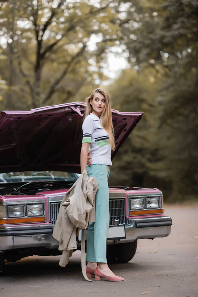 young woman holding cape and looking away while opening hood of broken vintage car - Fotografie, Obrázek