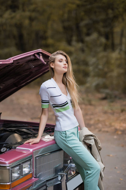 νεαρή κομψή γυναίκα κοιτάζει μακριά, ενώ στέκεται κοντά σε σπασμένο ρετρό αυτοκίνητο με ανοιχτή κουκούλα - Φωτογραφία, εικόνα
