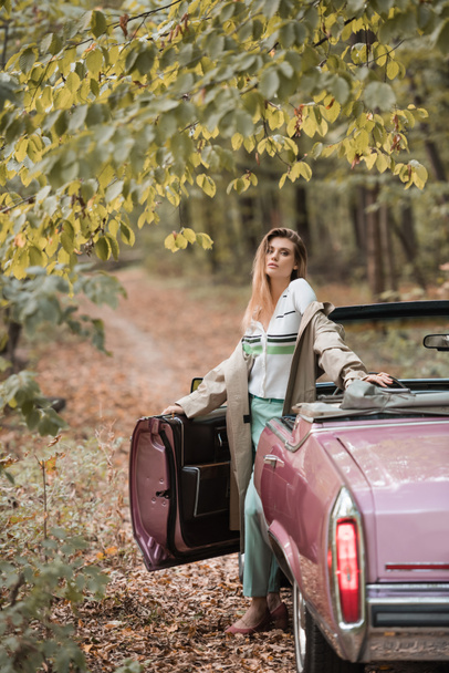 young stylish woman in cape looking at camera while standing near vintage cabriolet in forest - Foto, afbeelding