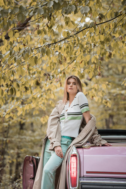 young woman looking at camera while standing near vintage cabriolet in forest - Photo, Image