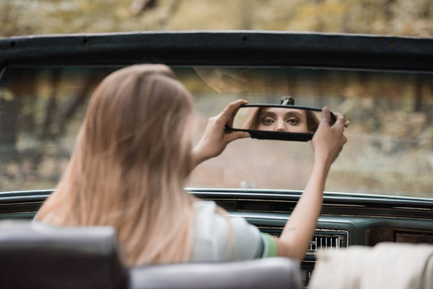 οπίσθια όψη νεαρής γυναίκας που προσαρμόζεται σε κάτοπτρο οδήγησης σε καμπριολέ σε θολή πρόσθια όψη - Φωτογραφία, εικόνα