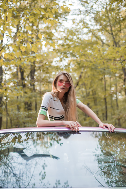 stylowa kobieta w okularach przeciwsłonecznych patrząca w kamerę opierając się na przedniej szybie kabrioletu - Zdjęcie, obraz