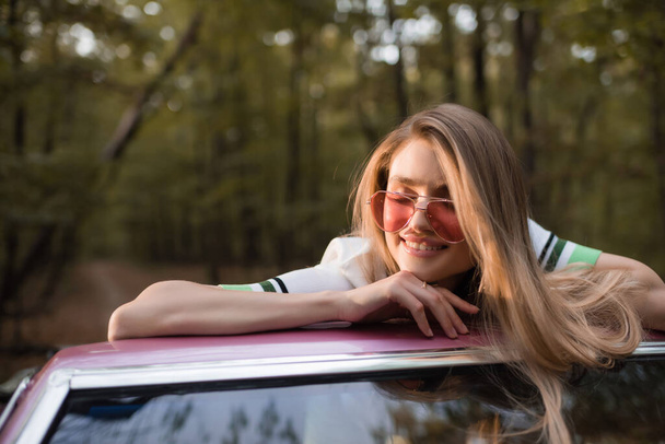 jeune femme en lunettes de soleil souriant les yeux fermés tout en s'appuyant sur le pare-brise de cabriolet - Photo, image