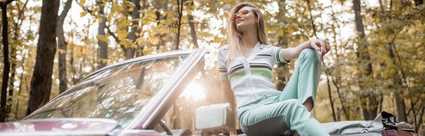 stylish woman in sunglasses looking away while posing in retro convertible car in sunshine, banner - Foto, Imagen