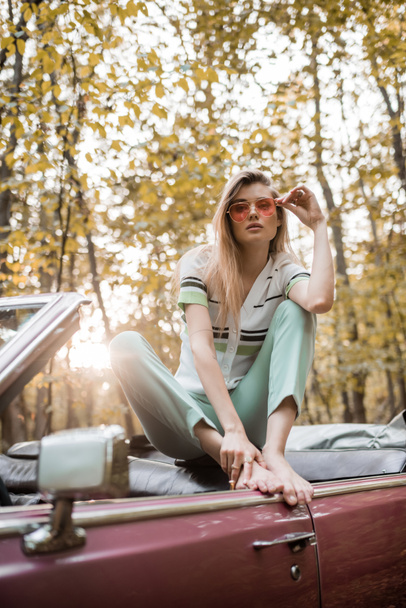 mujer descalza elegante tocando gafas de sol mientras posa en cabriolet en el bosque en primer plano borroso - Foto, imagen