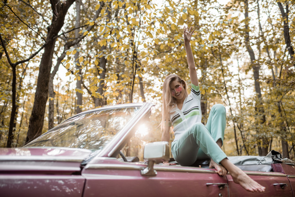 joyful barefoot woman looking at camera while posing with hand in air in cabriolet - Foto, Bild
