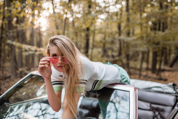stijlvolle jonge vrouw aanraken zonnebril terwijl leunend op voorruit van cabriolet - Foto, afbeelding