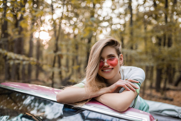 radostná žena v slunečních brýlích při pohledu na kameru, opírající se o čelní sklo kabrioletu se zkříženýma rukama - Fotografie, Obrázek
