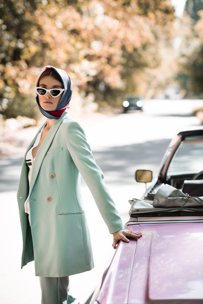 trendy woman in kerchief and sunglasses standing on road near cabriolet - Фото, изображение