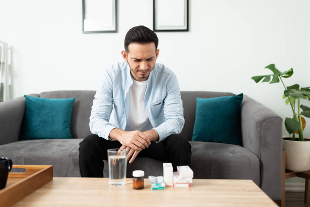 Привлекательный латиноамериканец сидит на диване и смотрит на кучу таблеток, которые ему прописал врач. - Фото, изображение