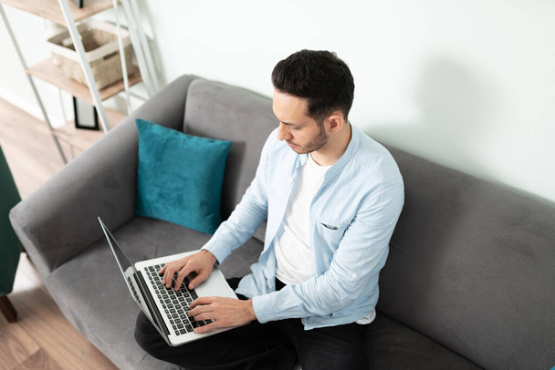Vista de ángulo alto de un hombre de 30 años sentado en su sofá de la sala de estar mientras trabajaba en su computadora portátil - Foto, Imagen