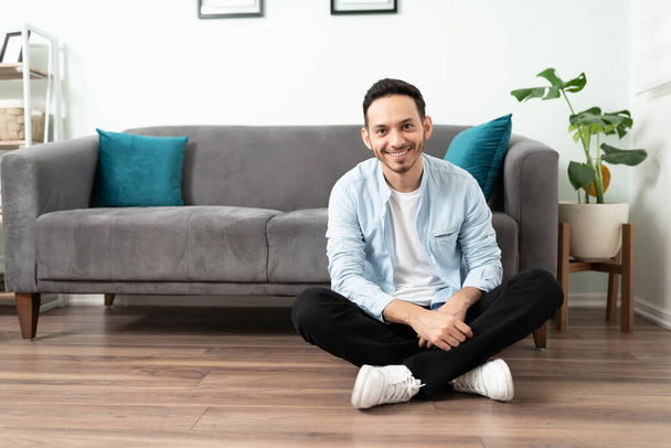 Handsome Hispanic man in his 30s smiling and looking confortable sitting on the floor at home - Photo, image