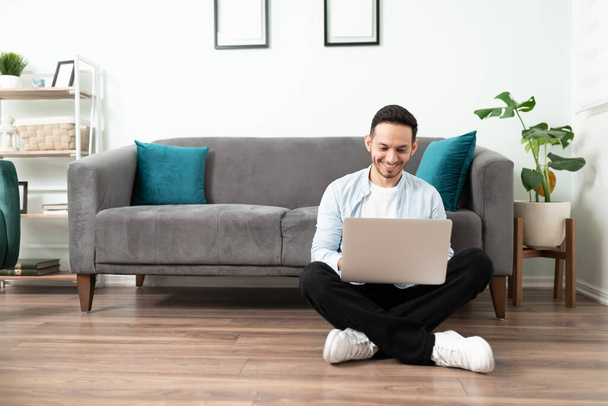 Όμορφος άνδρας κάθεται στο πάτωμα στο σπίτι και κάνει κάποια δουλειά από το σπίτι με έναν φορητό υπολογιστή - Φωτογραφία, εικόνα
