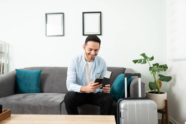 Красивый молодой человек, использующий приложение, чтобы поймать такси, сидя в своей гостиной диван рядом с чемоданом, готовый к путешествию - Фото, изображение