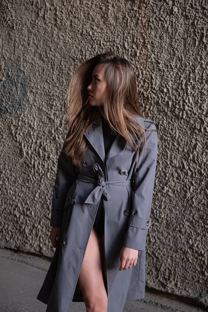 attractive young asian model in gray trench coat standing near wall outdoors - Valokuva, kuva