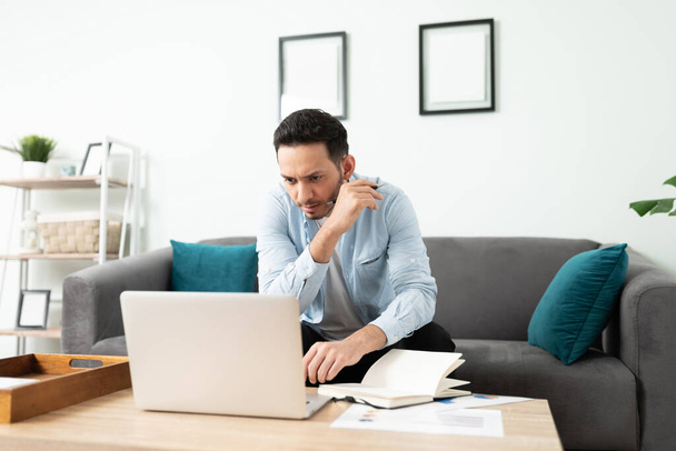 Латиноамериканец лет 30 выглядит занятым и сосредоточенным во время работы над ноутбуком из дома - Фото, изображение