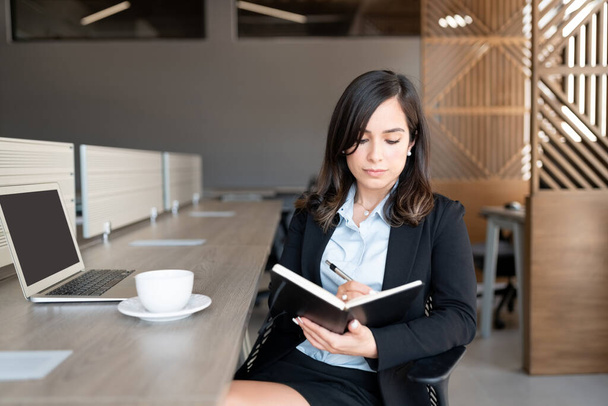 Προσωπογραφία της νεαρής γυναίκας που κάθεται στο γραφείο της κρατώντας σημειώσεις στο ημερολόγιο του γραφείου - Φωτογραφία, εικόνα
