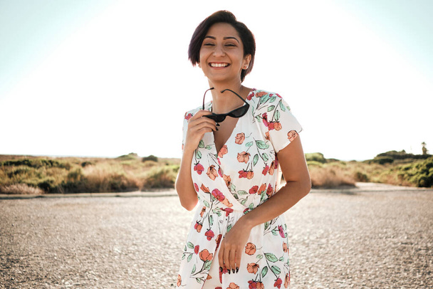 Portrait d'une belle fille latine souriante portant une robe décorée de fleurs. Elle est avec le soleil à son dos et porte des lunettes de soleil. - Photo, image