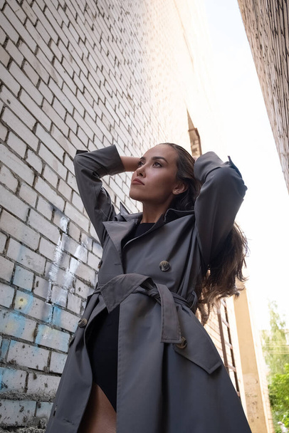 attraktives junges asiatisches Model im grauen Trenchcoat, das an der Wand im Freien steht - Foto, Bild